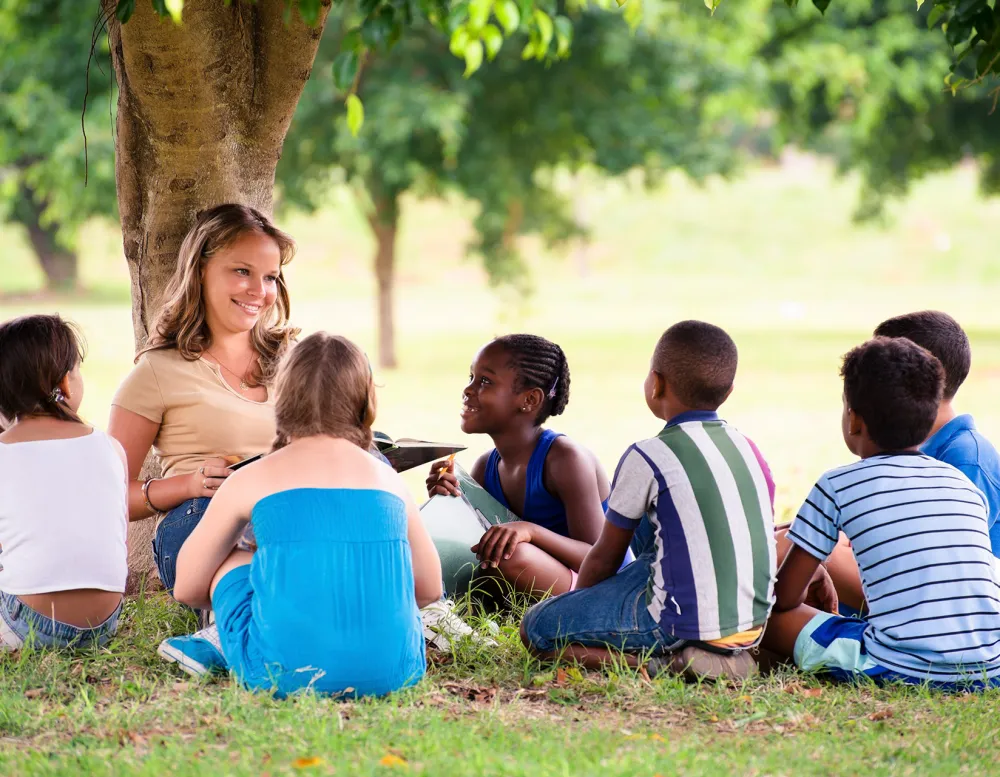 elementary students and teacher sitting under a tree.