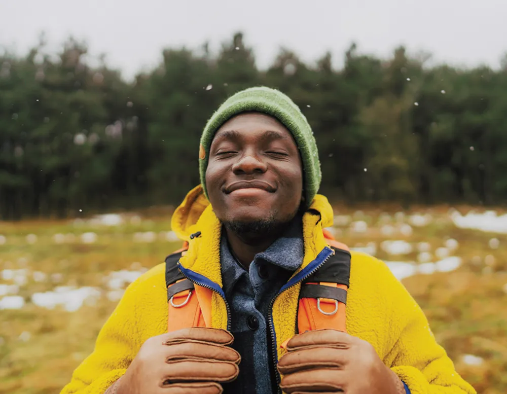 Man smiling with eyes closed, standing outside in front of rows of evergreen trees.