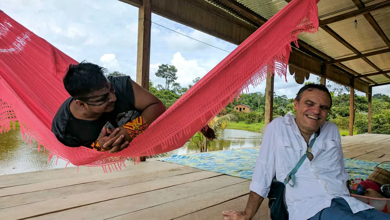 Two men laughing together next to a river, with one man in a red hammock.