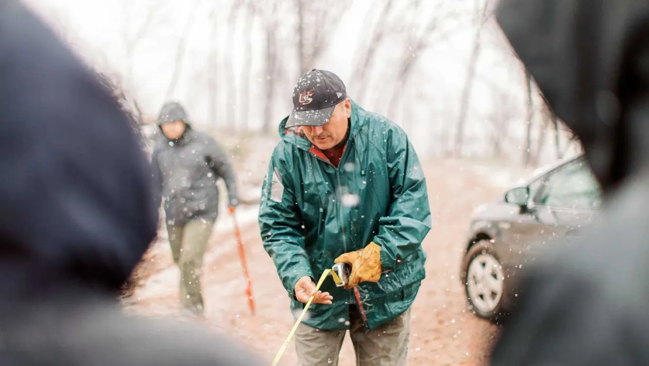 A man in a green jacket holds a measuring tape in a snowy landscape.