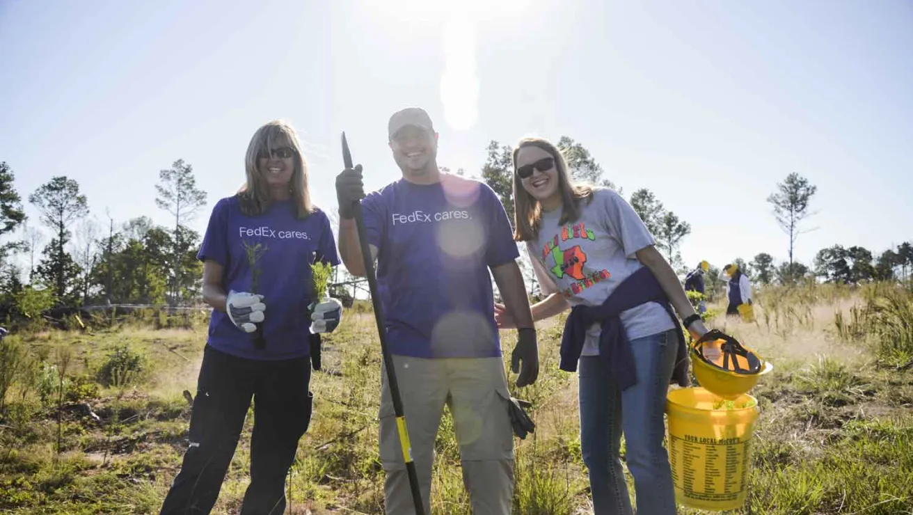 Three individuals in purple shirts stand together, each holding a shovel and a bucket, ready for planting trees.