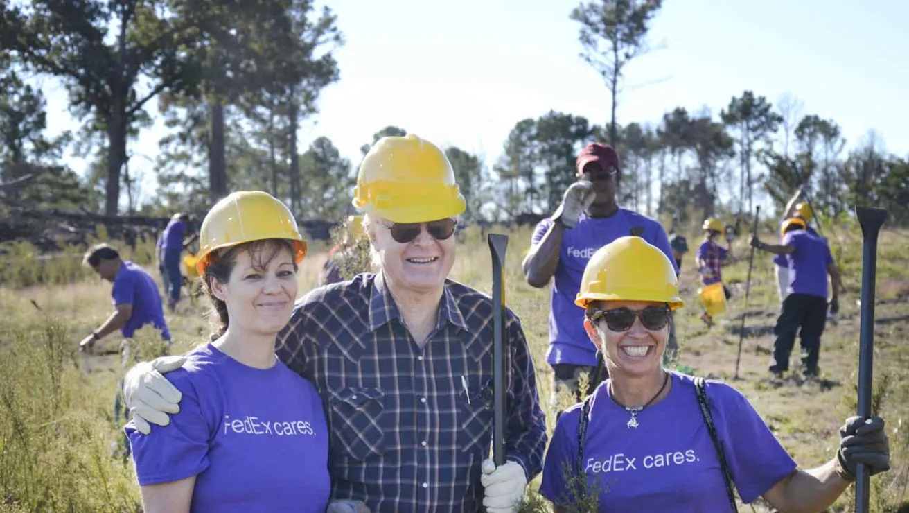 Three individuals wearing purple shirts and hard hats are standing together in a grassy field.