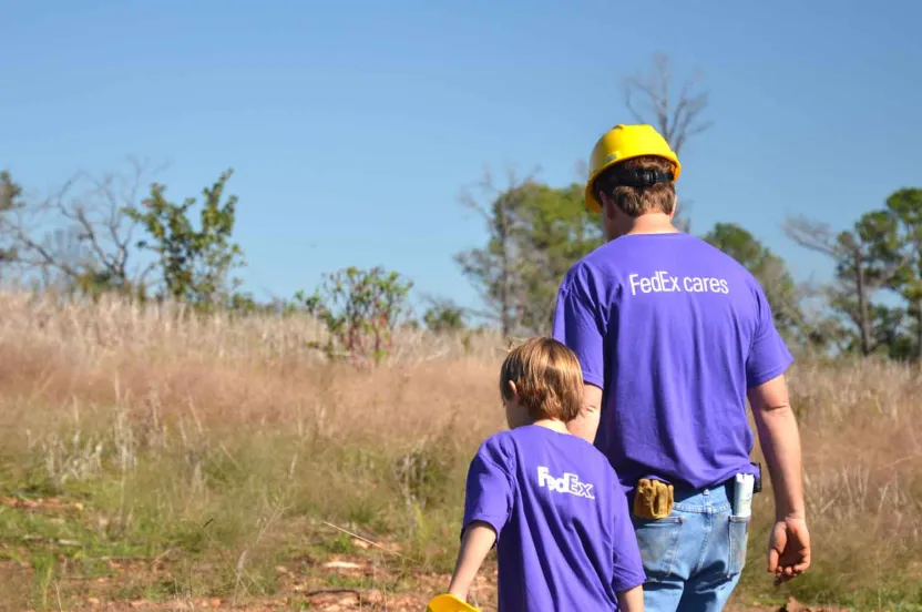 A man in yellow hard hat and blue FedEx t-shirt walks with a young boy in a forest.