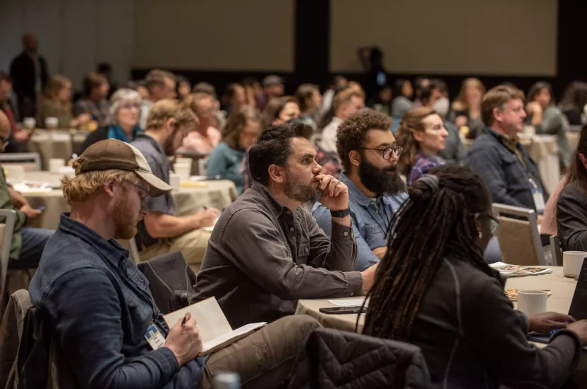 Several people sitting around tables at a conference listening to a speaker.