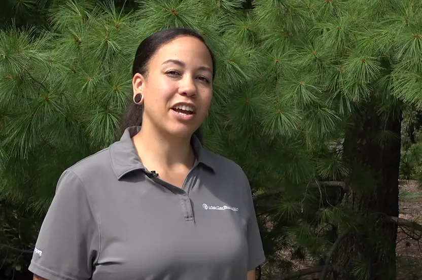 Woman talking to a camera in front of an eastern white pine tree.